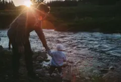 A imagem mostra um homem inclinado à beira de um riacho, segurando a mão de uma criança pequena que está de pé sobre pedras próximas à água. A cena é iluminada pelo sol baixo no horizonte, que cria um forte contraluz e reflexos alaranjados na lente. O homem veste camisa xadrez e calça escura, enquanto a criança usa um gorro claro e um casaco de tricô azul. Ao fundo, há um campo verde e uma fileira de árvores em silhueta, compondo uma paisagem rural tranquila ao entardecer.