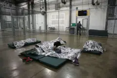 Una niña centroamericana sobre un colchón y una sábana térmica plateada en el suelo, junto a otros dos menores tumbados y cubiertos, en un espacio rodeado por una verja, cuya puerta cierra un agente, en un centro de detención de la Patrulla Fronteriza de Estados Unidos en McAllen, Texas, el 8 de septiembre de 2014. (Foto: John Moore/Getty Images)