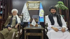 محمد اکبری، معاون شورای علمای شیعه افغانستان در دفتر والی بامیان، در کنار گل‌حیدر شفق نشسته است.
