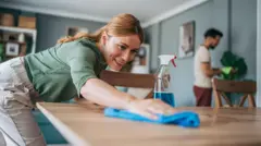 Mujer pasándole el trapo a la mesa con una sonrisa. 