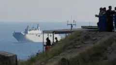 Kıyıdaki bir yükseltide insan silüetleri görünüyor ileride denizde bir donanma gemisi var. 