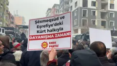 Kalabalığı arkadan gören bir fotoğrafta bir kişinin elinde Emek Partisi'nin Suriye'deki Kürt mahalleleriyle dayanışmayı vurgulayan bir pankartı görülüyor.