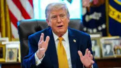 ダークスーツ、白シャツ、黄色系ネクタイを着けたトランプ氏がいすに座り、両手を前に出して口を開いている