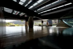 Interior sombreado con techo estructural visible, grandes vigas y columnas de piedra, abierto hacia un paisaje exterior iluminado.