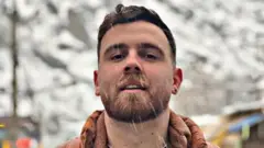 Um homem de cabelo escuro, barba e brinco na orelha olha para a câmera enquanto a neve cai ao seu redor