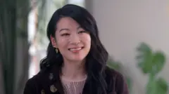 Arden Cho sorri para o lado da câmera. Ela tem cabelo castanho longo e ondulado e usa vários brincos de ouro, blusa creme e casaco marrom, em uma sala com paredes creme e uma janela atrás dela.