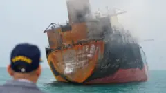 A man looks on as a rusting cargo ship catches fire in Iraqi territorial waters
