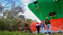 Três pessoas, vestindo jeans e jaquetas casuais, estão de pé, observando um enorme navio porta-contêineres verde que encalhou em terra firme. Uma casa parece pequena e está ao lado do navio.