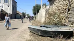 A small boat rests by the side of a road in Kherson, Ukraine, as residents carry bottled water