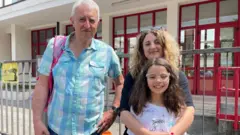 Um homem mais velho, uma mãe e uma filha são vistos em frente a um prédio