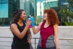 Duas mulheres em roupa de treino se olham rindo e fazem um brinde com duas bebidas isotônicas.