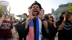 One Harvard University student dey with fellow demonstrators during one protest in support of international students for Harvard University. E wear graduation cap and gown wit sash wey get American flag.
