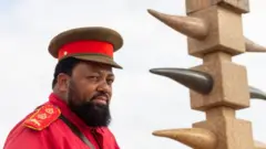 Un chef de haut rang portant des vêtements traditionnels herero se tient à côté d'un monument en l'honneur des OvaHerero