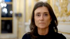 Sandrine Josso olha fixamente para a câmera. Ela tem cabelos escuros de comprimento médio e veste uma blusa preta. Está diante de uma parede decorada com folhas de ouro.