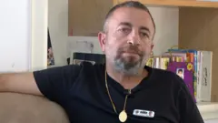 A close-up of a man in a black T-shirt sitting on a sofa. He has short dark hair, a short moustache and a short beard which are all flecked with grey. He is wearing a gold medallion.