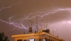 سماء مظلمة مليئة بالبرق فوق مبنى سكني يظهر من الأعلى، نافذتان مضاءتان في المبنى، وبقربه شجرة خضراء، في العاصمة الأردنية عمّان.