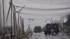 Мокра і брудна дорога з військовою технікою, накрита пошматованими білими сітками. Дорогою, повз танк із протидроновим "мангалом", йдуть двоє цивільних із візком і собакою. 