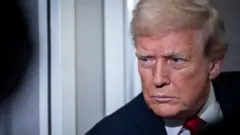 大統領専用機の通路の壁を背に、厳しい顔で前にいる記者団を見ているトランプ氏