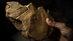 Un primer plano de un trozo roto de piedra de color marrón claro. Son claramente visibles los restos de algunas de las letras que quedaron impresas en el yeso al secarse: R-L-O-N. Significa Procurador de Londinium y muestra que el edificio del que formaba parte era un edificio civil en la ciudad en crecimiento.