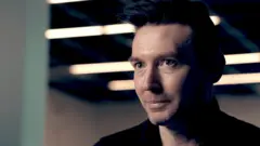 Matt Motyl, um homem de cabelo castanho penteado para trás, aparece vestindo uma camisa preta enquanto está sentado em um espaço com aparência industrial, com luzes lineares no teto visíveis ao fundo, desfocadas