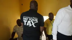 One man dey wear T-Shirt wit inscription for back "Pay Your Tax" dey walk down di stairs