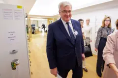 Сибіга у темному костюмі , білій сорочці і синій краватці йде поміж людьми. Посміхається і дивиться прямо 