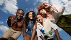 四位朋友靠在一起向下看著鏡頭，鏡頭從地面向上拍攝。他們都面帶笑容。兩名男子穿著短褲，一人搭配背心與頸巾，另一人穿著印花襯衫。一名女子穿牛仔連身短褲，另一名則穿綠色印花長裙與白色交叉上衣。他們在戶外，頭頂是藍天與零散白雲。