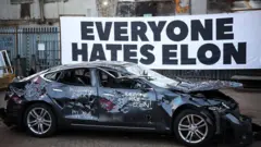 Carro danificado e pichado na frente de cartaz que diz: 'Todos odeiam Elon'
