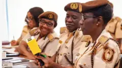 Aworan yii ṣafihan awọn osiṣẹ ileeṣẹ to n ri si iwọle-jade awọn arinrinajo lorilẹede yii ( Nigeria Immigration Servive). Mẹrin ni awọn oṣiṣẹ naa ti wọn wọṣọ iṣẹ wọn, obinrin mẹta ati ọkunrin kan. Oṣiṣẹ-binrin to wa lapa ọtun mu ontẹ ti wọn fi n lu iwe dani, awon ẹlẹgbẹ rẹ yoku n wo o. 