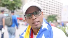 Un hombre con gorra blanca y la bandera de Venezuela en los hombros mira a cámara.