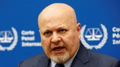Прокурор Международного уголовного суда Карим Хан выступает во время интервью, сидя перед синей табличкой с размытыми белыми надписями и символом МУС — весами правосудия, окруженными лавровым венком.