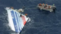 白地に赤と青の複数の線が斜めに入った旅客機の機体の一部が、海上に浮かんでいる。オレンジのライフジャケットを着た人たちが乗る灰色のゴムボートからけん引用のロープが伸び、ライフジャケットを着た人たちが水中に入り、機体を手でつかんで引いている