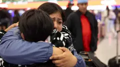 A imigrante equatoriana Andrea, de 28 anos, recebe um longo abraço da sua irmã, de frente para a câmera, antes do seu voo para o Equador no Aeroporto Internacional JFK em Nova York, no dia 26 de outubro de 2025. Andrea e sua prima decidiram se "autodeportar" com seus filhos, depois que seus maridos foram detidos por agentes do Serviço de Imigração e Alfândega dos Estados Unidos (ICE) e deportados para o Equador, por não terem documentos. A filha mais nova de Andrea, Shanell, de 7 meses, nasceu nos Estados Unidos e é cidadã americana.