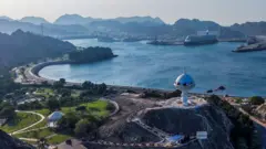 نمایی هوایی از کورنیش مطرح در مسقط، عمان. جاده‌ای ساحلی و منحنی در امتداد آب‌های آبی‌رنگ خلیج کشیده شده و فضاهای سبز در کنار آن دیده می‌شود. در پیش‌زمینه، سازه‌ای سفید با گنبد آبی‌رنگ بر فراز تپه‌ای سنگی مشرف به بندر قرار دارد. در پس‌زمینه، کوه‌ها و چند کشتی بزرگ تفریحی که در ساحل پهلو گرفته‌اند، زیر آسمانی صاف دیده می‌شوند.