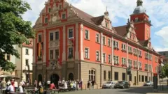 A imagem mostra um edifício histórico de fachada vermelha e detalhes ornamentais, com uma torre e relógio, localizado em uma área urbana movimentada por pedestres e veículos.