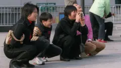 Un grupo de personas en un espacio público descansan en cuclillas profundas mientras se ríen. Están en equilibrio cerca del suelo con las rodillas completamente flexionadas y los pies apoyados en el suelo. Uno tiene una cámara en la mano y está tomando fotografías. Al fondo, a la derecha, hay otro grupo de personas que están de pie.