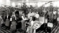 A imagem em preto e branco mostra o interior de uma fábrica no início do século 20, com dezenas de trabalhadores distribuídos entre grandes máquinas industriais. Muitos usam aventais e bonés, enquanto operam equipamentos ou posam para a fotografia. O ambiente é amplo, iluminado por janelas altas, e transmite o ritmo intenso da produção industrial da época.