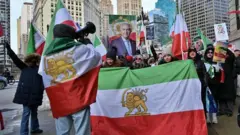 Ellerinde ve üzerlerinde aslan ve güneş motifli İran bayrakları taşıyan muhalif göstericiler eylem yapıyor. Bir tanesinin elinde megafon var ve kalabalığa seslendiği görülüyor.