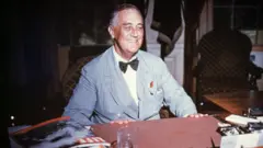 Retrato de Franklin Delano Roosevelt sentado em sua mesa, sorrindo.