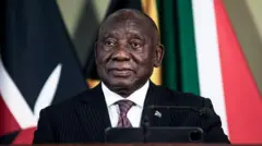 South Africa President, Cyril Ramaphosa wear black suit, brown neck-tie wey get designs and white inner shirt. E sand for podium as e look inside di camera.