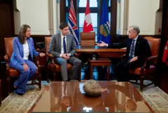 Смит, Эби и Карни сидят на креслах спиной к длинному столу, Смит и Эби с одной стороны (на фото слева), Карни с другой, на некотором расстоянии от них. За столом стоят флаги, за ними окно. На переднем плане еще один стол, в котором отражаются политики, на нем лежит сувенир в виде камня, на котором вырезаны извилистые линии.