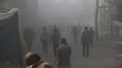 فلسطينيون يسيرون وسط الدخان والغبار بعد الهجمات الإسرائيلية على مبنى في مخيم البريج للاجئين في غزة