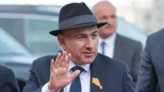 Paşinyan kadraj dışındakilere selam veriyor. Üzerinde takım elbise, kafasında fötr şapka var. Yakasında turuncu Ermenistan rozeti takılı.