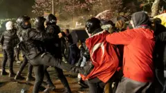 An armoured riot police officer can be seen kicking out at a man wearing a Turkish flag 