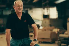 جون هارب ذو شعر رمادي قصير وشارب رمادي. يرتدي قميصاً أسود قصير الأكمام وبنطالاً أخضر، ويجلس في مصنع الطوب الخاص به، وخلفه صناديق مفتوحة.