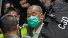 Jimmy Lai wears a green face mask and arrives to a crowded court room, he is seen outside of the courtroom with security around him.