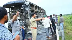 The charred remains of the bus that caught fire, with several people, including a police officer in uniform, standing nearby. 