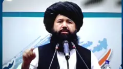 وزیر تحصیلات عالی حکومت طالبان با لنگی و واسکت سیاه و پیراهن سفید پشت مایک در حال صحبت کردن است.