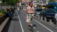 Eetek ve tşört giyinmiş bir kadın güneş gözlükleri takmış, e-scooter yolundan gidiyor, yan tarafta trafik akıyor, diğer yanı yayalara ayrılmış. 