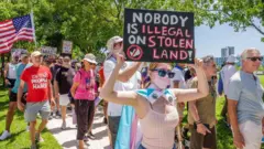 Manifestantes y una joven en primer plano con una pancarta que dice: "Nadie es ilegal en una tierra robada"